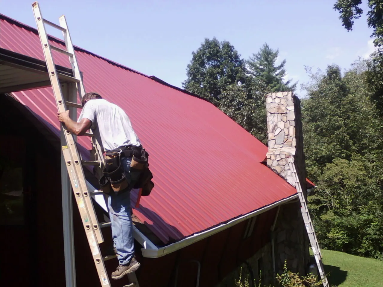 Roofer on a ladder beside a red metal roof and stone chimney — Soffit & Fascia Repair by Roofing Brothers in Blythe